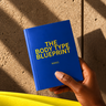 A hand holding a blue book titled 'THE BODY TYPE BLUEPRINT' by Nikko, set against a textured concrete wall with diagonal shadows and a bright yellow foreground element.
