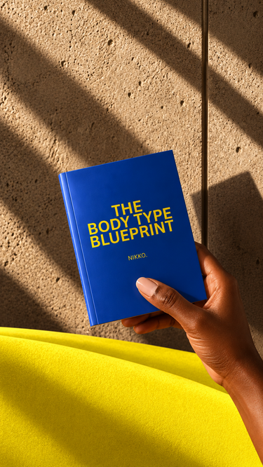 A hand holding a blue book titled 'THE BODY TYPE BLUEPRINT' by Nikko, set against a textured concrete wall with diagonal shadows and a bright yellow foreground element.
